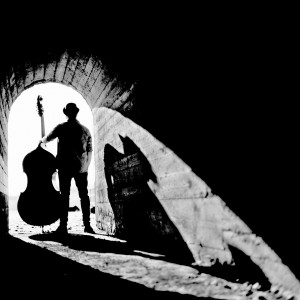 Adam Ben Ezra holding his double bass standing back at the end of a tunnel with his shadow.