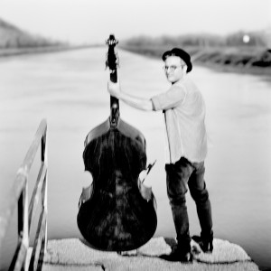 Adam Ben Ezra with his double bass standing at a river looking to the photo camera.