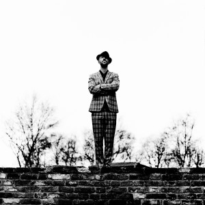 Charlie Winston standing on old brick wall with trees in the background wearing suit and a hat.