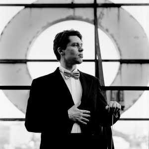 Kevin Griffiths holding baton in his hand wearing tuxedo in front of a giant letter O with a city view in the background.