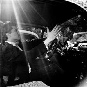 Kevin Griffiths making a gesture with his hand inside an old car wearing a tuxedo.