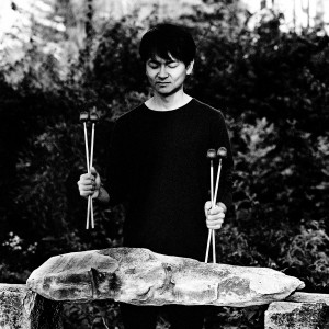 Masayoshi Fujita standing at his vibraphone holding the sticks in front of trees.